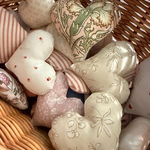 Puede incluir: Una cesta de mimbre llena de varios cojines de tela en forma de corazón. Los cojines están hechos de diferentes telas con patrones florales y geométricos en tonos de rosa, blanco y verde.