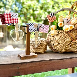 May include: A wooden stand with two heart-shaped glass vases holding small flags made of fabric with red, white, and blue patterns. The stand is on a wooden table with a wicker basket filled with yellow flowers and a small wicker box.