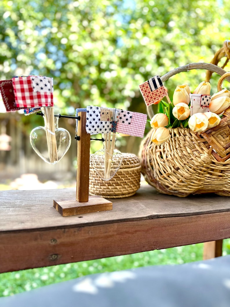 May include: A wooden stand with two heart-shaped glass vases holding small flags made of fabric with red, white, and blue patterns. The stand is on a wooden table with a wicker basket filled with yellow flowers and a small wicker box.