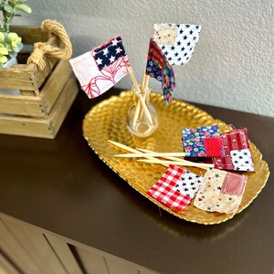 May include: A gold tray with six small fabric flags on wooden skewers. The flags are made of red, white, and blue patchwork fabric with stars and other patterns. The flags are arranged in a circle on the tray.