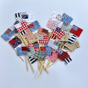 Könnte beinhalten: Eine Sammlung von 20 kleinen Stoff-Flaggen mit roten, weißen und blauen Patchwork-Designs. Die Flaggen sind an Holzspießen befestigt und eignen sich perfekt zum Dekorieren von Kuchen, Cupcakes oder anderen Desserts.