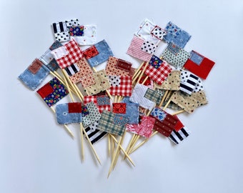 May include: A collection of 20 small fabric flags with red, white, and blue patchwork designs. The flags are attached to wooden skewers and are perfect for decorating cakes, cupcakes, or other desserts.