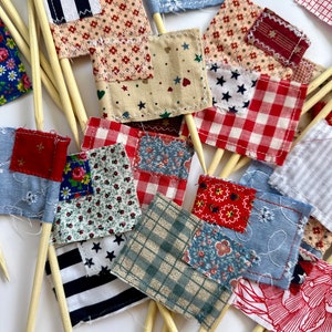 May include: A collection of small fabric flags with various patterns, including red and white gingham, blue and white stripes, and floral prints. The flags are attached to wooden skewers.
