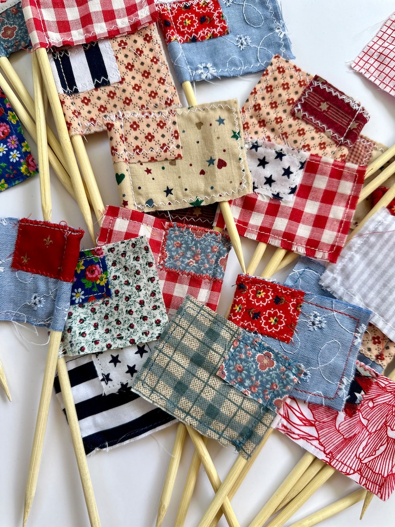 May include: A collection of small fabric flags with various patterns, including red and white gingham, blue and white stripes, and floral prints. The flags are attached to wooden skewers.