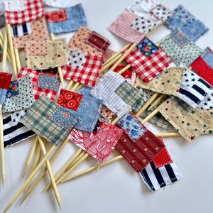 May include: A collection of small fabric flags with red, white, and blue patchwork designs. The flags are attached to wooden skewers, perfect for decorating cakes or cupcakes.
