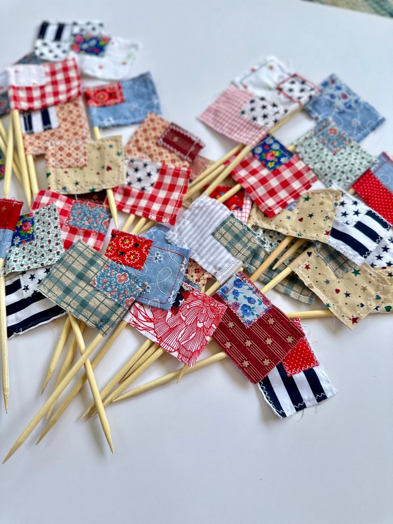 May include: A collection of small fabric flags with red, white, and blue patchwork designs. The flags are attached to wooden skewers, perfect for decorating cakes or cupcakes.