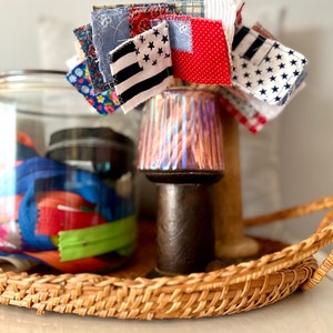 May include: A wicker tray with a glass jar filled with colorful fabric scraps and a small lamp with a colorful patterned shade. A cluster of small fabric squares with stars and stripes patterns are attached to the lamp.
