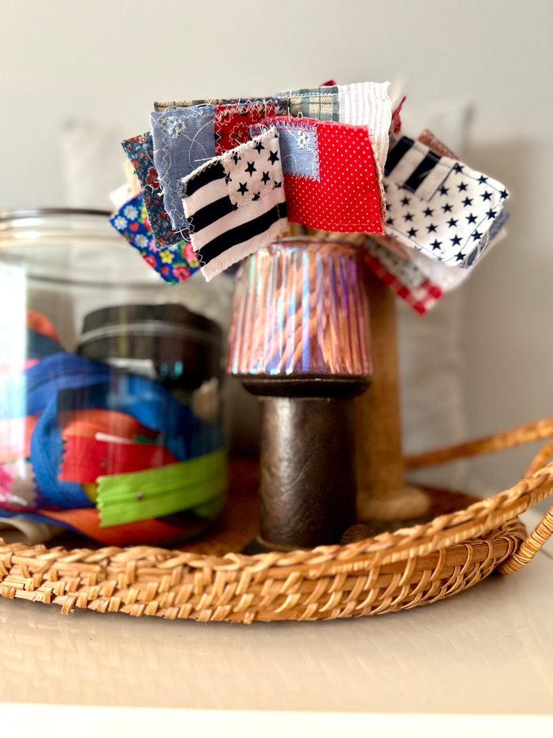 May include: A wicker tray with a glass jar filled with colorful fabric scraps and a small lamp with a colorful patterned shade. A cluster of small fabric squares with stars and stripes patterns are attached to the lamp.