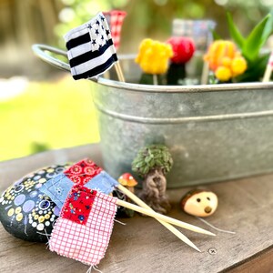May include: A small galvanized metal bucket filled with miniature flags, flowers, and a small tree. The bucket is sitting on a wooden surface with a painted rock and two wooden skewers with fabric flags.