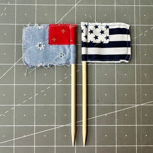 May include: Two small fabric flags on wooden skewers. The flag on the left is blue, red, and white with a fleur-de-lis design. The flag on the right is a blue and white striped flag with white stars.