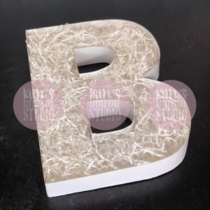 Pode incluir: Uma grande letra "B" branca preenchida com papel picado. A letra &eacute; feita de um material espesso e resistente e tem um design limpo e moderno. O fundo &eacute; escuro, fazendo a letra branca se destacar.