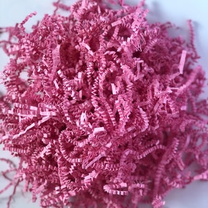 May include: A close-up shot of a pile of pink shredded paper. The paper is cut into thin strips and has a crinkled texture. The paper is a light, pastel pink color and is bunched together in a loose, irregular shape. This is often used as packaging material.