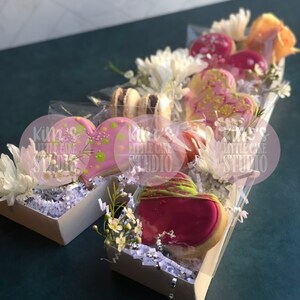 Pode incluir: Um arranjo em caixa de presente com biscoitos em forma de cora&ccedil;&atilde;o com cobertura rosa e granulado decorativo. A caixa tamb&eacute;m cont&eacute;m macarons, cake pops e flores brancas. O texto "kim's LITTLE CAKE STUDIO" &eacute; vis&iacute;vel.