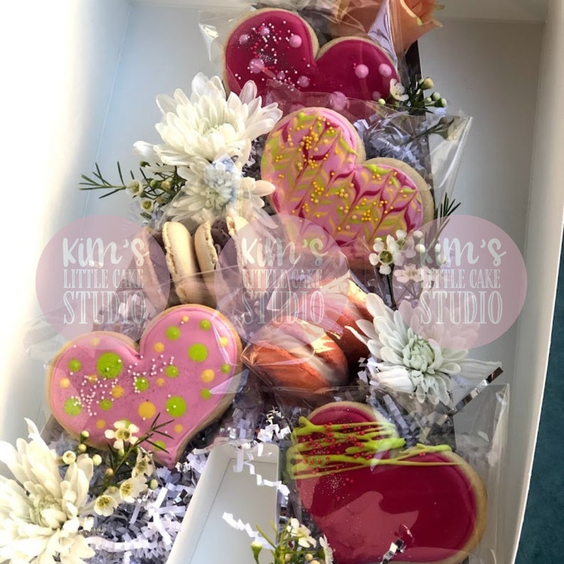 Pode incluir: Uma caixa de presente cheia de biscoitos em forma de cora&ccedil;&atilde;o, macarons e flores brancas. Os biscoitos s&atilde;o decorados com glac&ecirc; rosa, granulado e detalhes verdes e amarelos. A caixa &eacute; forrada com papel branco.