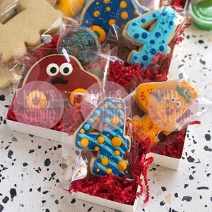 Pode incluir: Variedade de biscoitos de a&ccedil;&uacute;car decorados em embalagens transparentes, incluindo um carro vermelho, o n&uacute;mero 4 e formas de dinossauros. Os biscoitos s&atilde;o exibidos em caixas brancas com papel de seda vermelho. Um pequeno recipiente de granulado tamb&eacute;m &eacute; vis&iacute;vel.