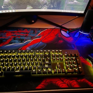 Op de afbeelding: Een zwart mechanisch toetsenbord met geel verlichte toetsen en een zwarte gamingmuis met groene verlichting, beide rustend op een grote rechthoekige bureaumat met een rode en blauwe Spider-Man grafische print op een donkere achtergrond.