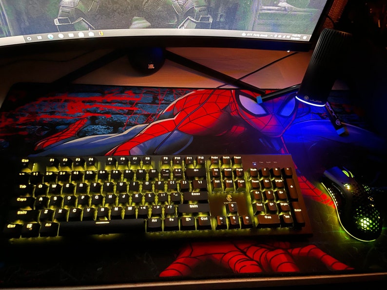 Op de afbeelding: Een zwart mechanisch toetsenbord met geel verlichte toetsen en een zwarte gamingmuis met groene verlichting, beide rustend op een grote rechthoekige bureaumat met een rode en blauwe Spider-Man grafische print op een donkere achtergrond.