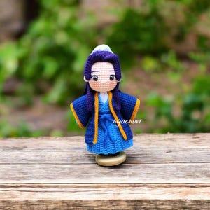 Puede incluir: Muñeca de ganchillo hecha a mano con un atuendo azul y amarillo. La muñeca tiene un sombrero blanco, pelo morado y tez clara. Está sobre una superficie de madera. El nombre de la muñeca es "ngochovi".