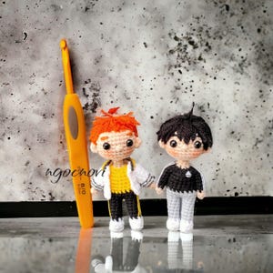 Può includere: Due bambole amigurumi all'uncinetto, una con i capelli arancioni e un vestito giallo e nero, l'altra con i capelli neri e un vestito nero e grigio. Le bambole sono in piedi su una superficie grigia.