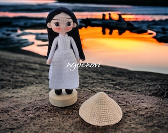 May include: A crocheted doll wearing a white dress and black shoes stands on a wooden base. The doll has long black hair and a sweet expression. A crocheted conical hat sits on the ground beside the doll. The background is a blurry image of a beach at sunset.