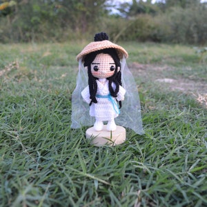 May include: A crocheted doll wearing a white dress, a straw hat, and a white veil. The doll is standing on a wooden base and is surrounded by green grass.