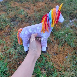 Puede incluir: Un juguete de unicornio de crochet con cuerpo morado, estrellas blancas y una melena arcoíris. El unicornio está siendo sostenido por una mano.