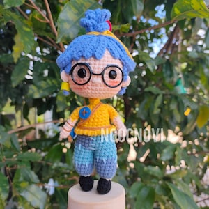 May include: A crocheted doll with blue hair, wearing yellow and blue clothing and glasses. The doll is standing on a wooden base.