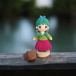 以下が含まれることがあります： 緑色の髪の毛、緑と黄色のドレス、ピンクのスカートを着た、かぎ針編みのドール。ドールは、小さな茶色のかぎ針編みのマグカップを持っています。ドールは木製の台座の上に立っています。