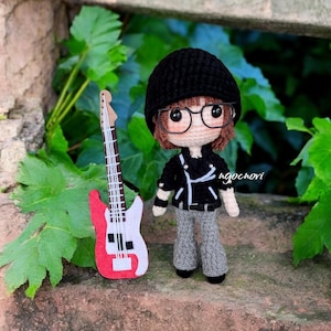 May include: A crocheted doll wearing a black beanie, glasses, and a black jacket with white accents. The doll is holding a red and white electric guitar. The doll is standing on a brown surface with green foliage in the background.