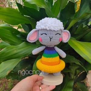 Può includere: Una bambola di pecora di uncinetto grigia che indossa un vestito arcobaleno con un galleggiante giallo all'uncinetto. La bambola è in piedi su una base bianca.