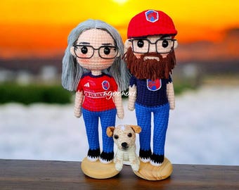 May include: Crocheted amigurumi figures of a couple with a dog. The woman has long gray hair, glasses, and is wearing a red shirt with blue jeans. The man has a red hat, glasses, a beard, and is wearing a blue shirt with blue jeans. The dog is white and brown and is sitting in front of the couple.