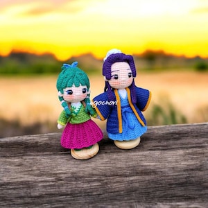 Puede incluir: Dos muñecas de ganchillo hechas a mano, una en verde y rosa, la otra en azul y morado, tomadas de la mano. Las muñecas tienen rasgos faciales detallados y visten ropa de estilo tradicional. El fondo es una puesta de sol borrosa sobre el agua.