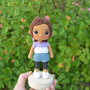 May include: Crocheted doll with brown hair, a purple and white striped shirt, blue denim shorts, and black leggings. The doll is wearing white shoes and is standing on a wooden base.