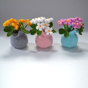 Puede incluir: Tres arreglos florales de ganchillo en macetas de diferentes colores. Las flores son blancas, amarillas y rosas. Las macetas son grises, rosas y azules.