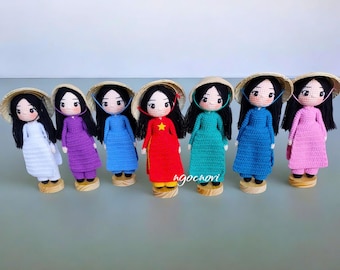 May include: A collection of seven handmade crochet dolls, each wearing a different colored dress and a traditional conical hat. The dolls have black hair and are standing on small wooden bases. The dresses are white, purple, blue, red, teal, blue, and pink.