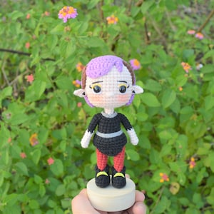 May include: A crocheted doll with purple hair, black and white clothing, and red leggings. The doll is standing on a wooden base and has a cartoonish appearance.