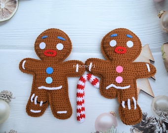 May include: Two crocheted gingerbread people holding a candy cane. The gingerbread people are brown with white icing details and colorful buttons.