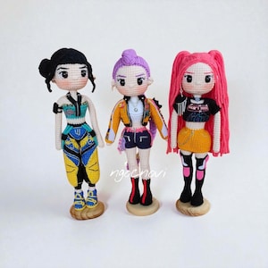 May include: Three handmade crochet dolls with detailed outfits and hairstyles. The dolls have black, purple, and pink hair, and wear colourful tops, jackets, shorts, and skirts. Each doll stands on a small wooden base.