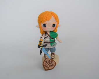 May include: A crocheted plush toy of Link from the Legend of Zelda video game series. The toy is wearing a green tunic, brown pants, and a yellow wig. The toy is holding a blue sword and is standing on a wooden base with a shield.