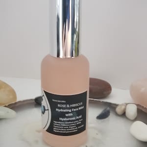 May include: A pink hydrating face mist bottle with a silver cap. The label reads "Rose & Hibiscus Hydrating Face Mist with Hyaluronic Acid." The bottle is surrounded by decorative stones.