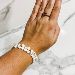 Op de afbeelding: Twee witte kralenarmbanden met gouden letters die "I Will Follow You" spellen.