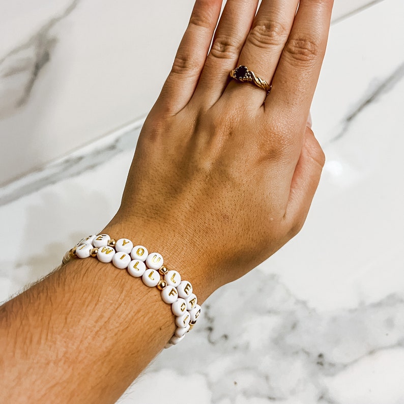 Op de afbeelding: Twee witte kralenarmbanden met gouden letters die "I Will Follow You" spellen.