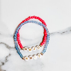 May include: Two beaded bracelets. One bracelet is red, the other is light blue. Both bracelets have white letter beads with gold accents. The top bracelet spells out "WWJD" and the bottom bracelet spells out "HWLF".