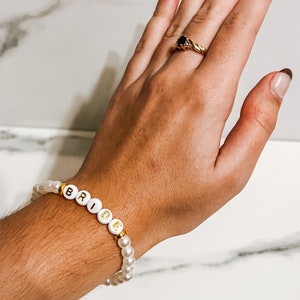 Pode incluir: Um bracelete de contas brancas com letras douradas que soletram "BRIDE".
