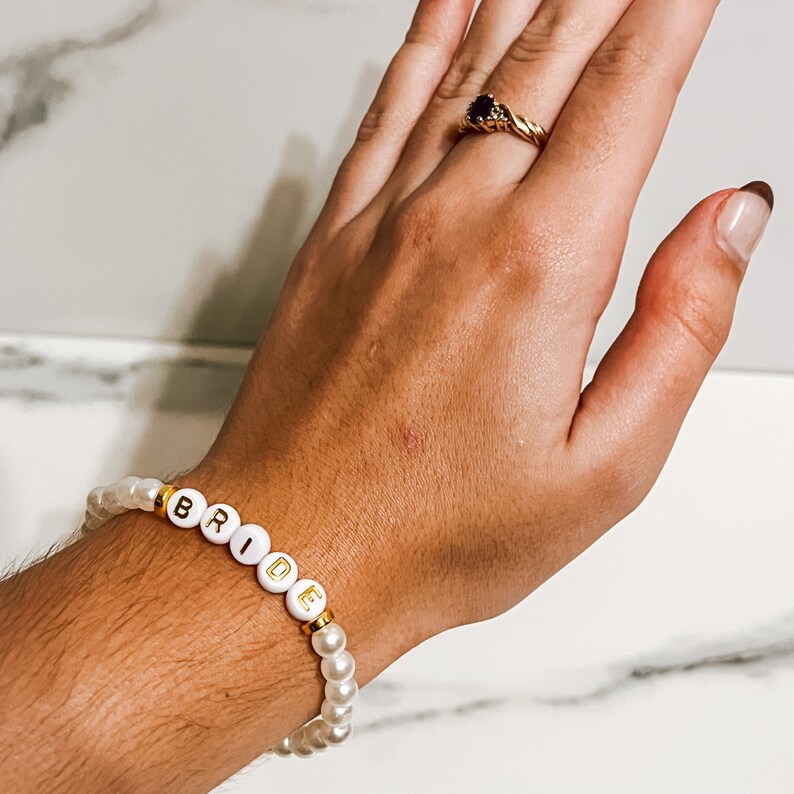 Pode incluir: Um bracelete de contas brancas com letras douradas que soletram "BRIDE".