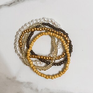 May include: A set of five beaded bracelets in white, brown, and gold. The bracelets are stacked on top of each other.