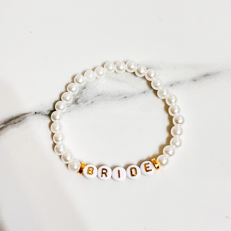 Pode incluir: Pulseira de p&eacute;rolas brancas com letras douradas que soletram "BRIDE".