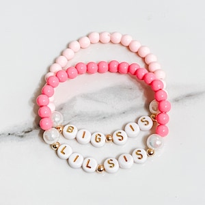 May include: Two pink and white beaded bracelets with gold letters spelling "Big Sis" and "Lil Sis".