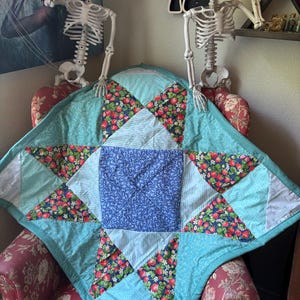 Pode incluir: Uma colcha de retalhos quadrada com um quadrado central de flores azuis rodeado por triângulos alternados de tecido com padrão de morangos sobre fundo azul escuro, tecido às riscas azul-claro e tecido azul-claro com desenhos de linhas brancas.