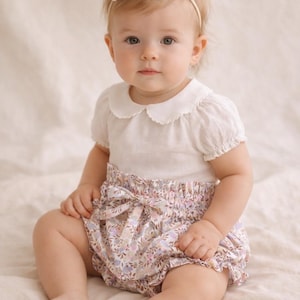 Puede incluir: Un bebé con una blusa blanca con cuello festoneado y mangas cortas, combinada con un bloomers con estampado floral. Una diadema con lazo a juego completa el conjunto. El bebé está sentado sobre una superficie de color claro.